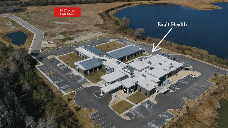 Aerial photograph of the completed Exalt Health Inpatient Rehabilitation Hospital campus and parking, labelled Exalt Health with an arrow, with the adjacent 14.87 acres marked for sale and the spring-fed lake to the right.